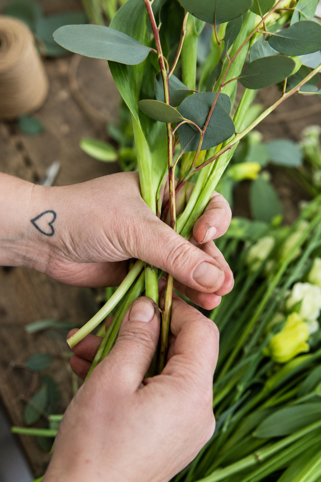 Hands bringing green flower stems and eucalyptus together while tying a bouquet
