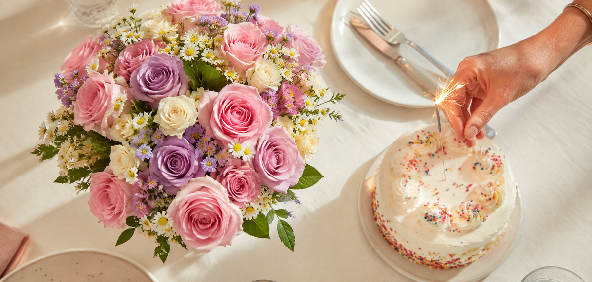 Pink and lilac rose bouquet beside a birthday cake and lit sparkler